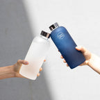 Two hands holding a clear water bottle and a blue water bottle with a brand logo against a gray background.
