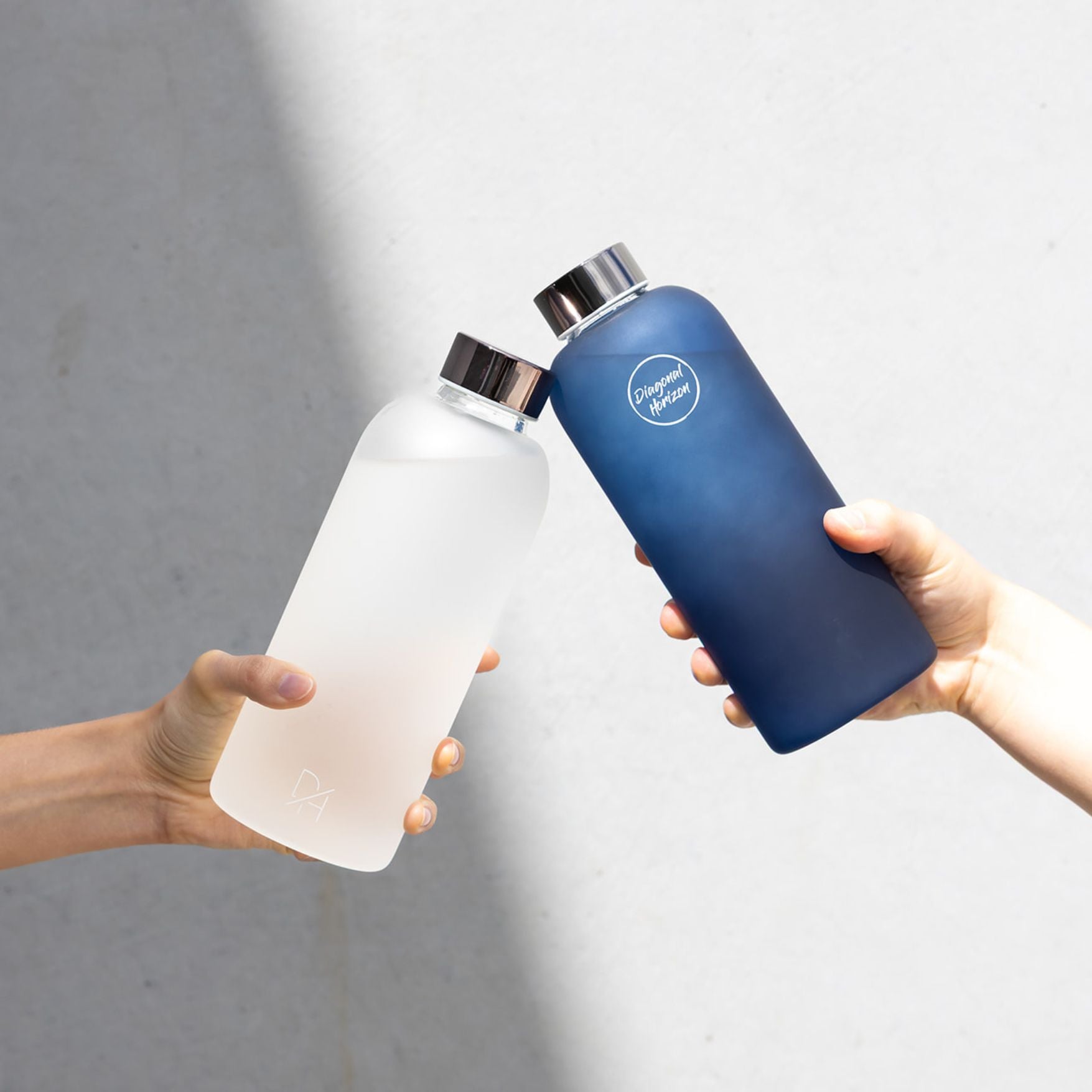 Two hands holding a clear water bottle and a blue water bottle with a brand logo against a gray background.