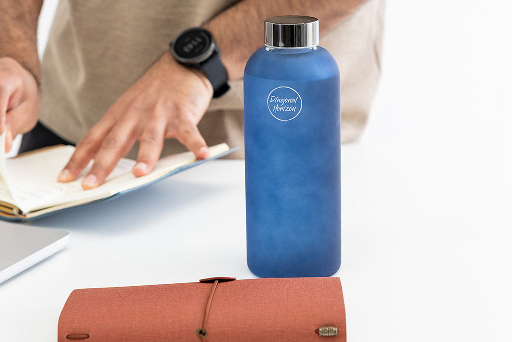 Blue glass frosted water bottle with a logo on a desk next to a person using journal.