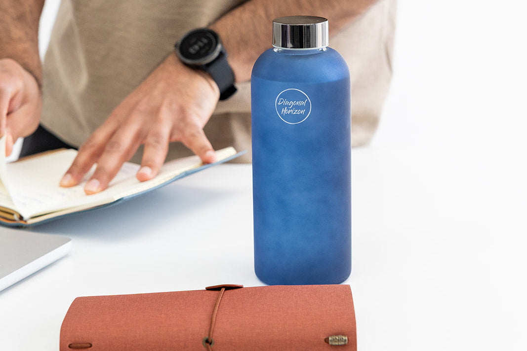 Blue glass frosted water bottle with a logo on a desk next to a person using journal.